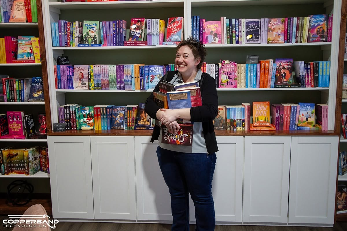 Liz Grant of Copperband holding an armload of books in front of the shelves at Blush and Bashful